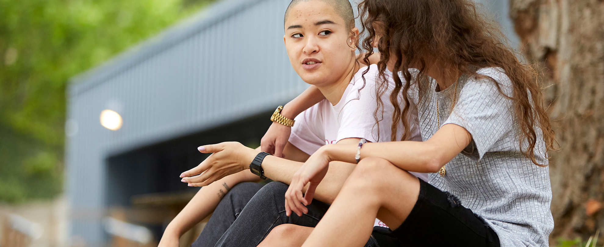 a-young-woman-with-curly-hair-and-in-grey-shirt-and-black-shorts-with-face-unseen-has-her-hands-wrapped-another-young-woman-with-shaved-hair-and-in-white-shirt-and-blakc-trousers-talking-to-her-while-they-sat-on-the-grass-with-a-tree-and-building-on-their-background
