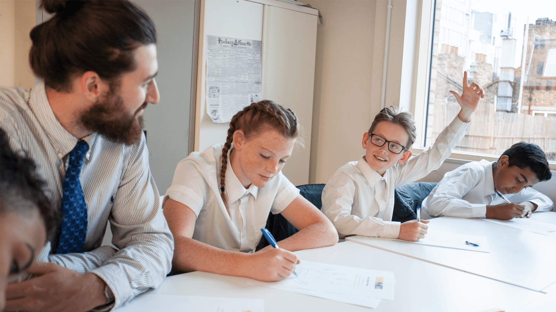 A male teacher sitting with his students at their desk smiling 
