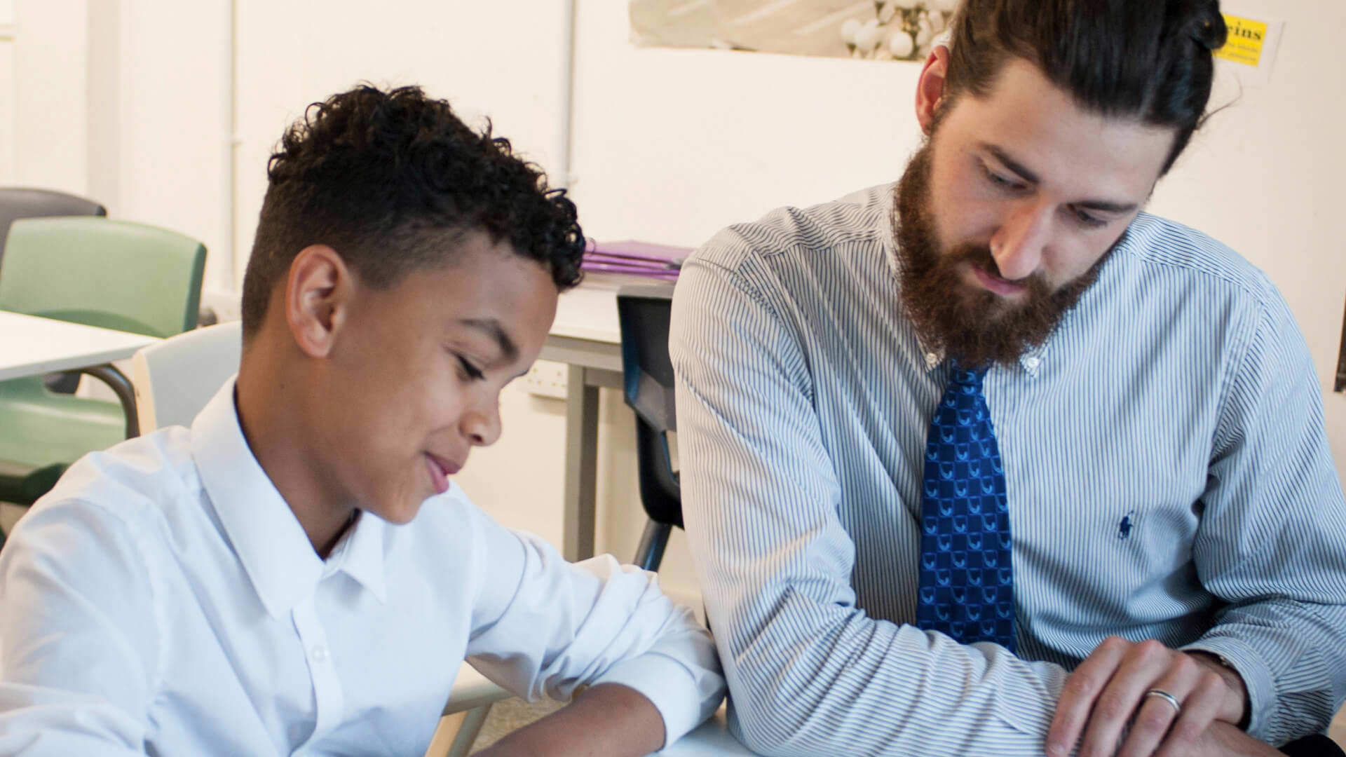 a teacher sits beside a student to help him read his lesson on his desk inside the classroom