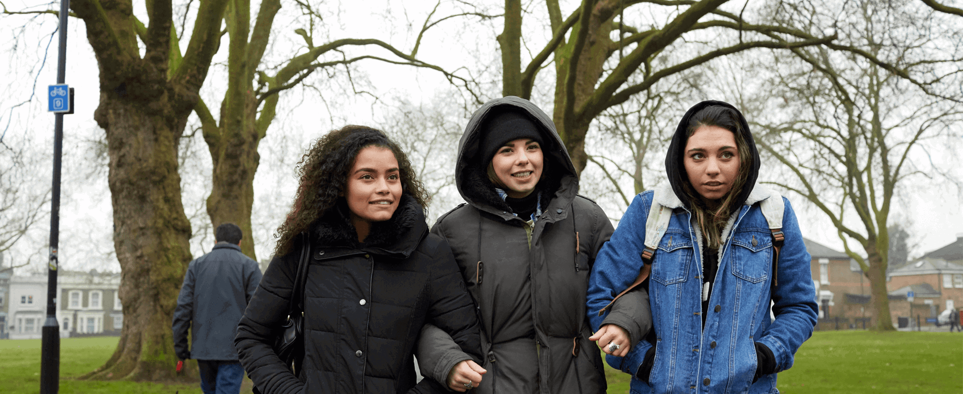 wide shot of three girls walking with linked arms on a tree lined street on a chilly day