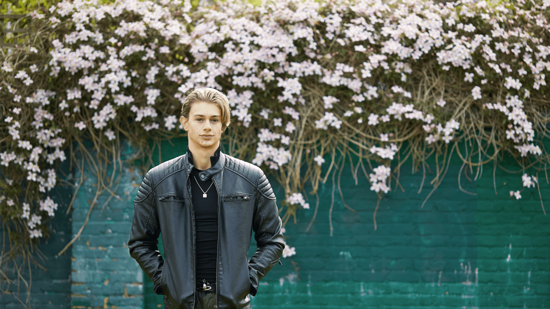 a boy wearing black jacket with hands on his pockets standing and looking in front of the camera on the backgound is a wall with flowers