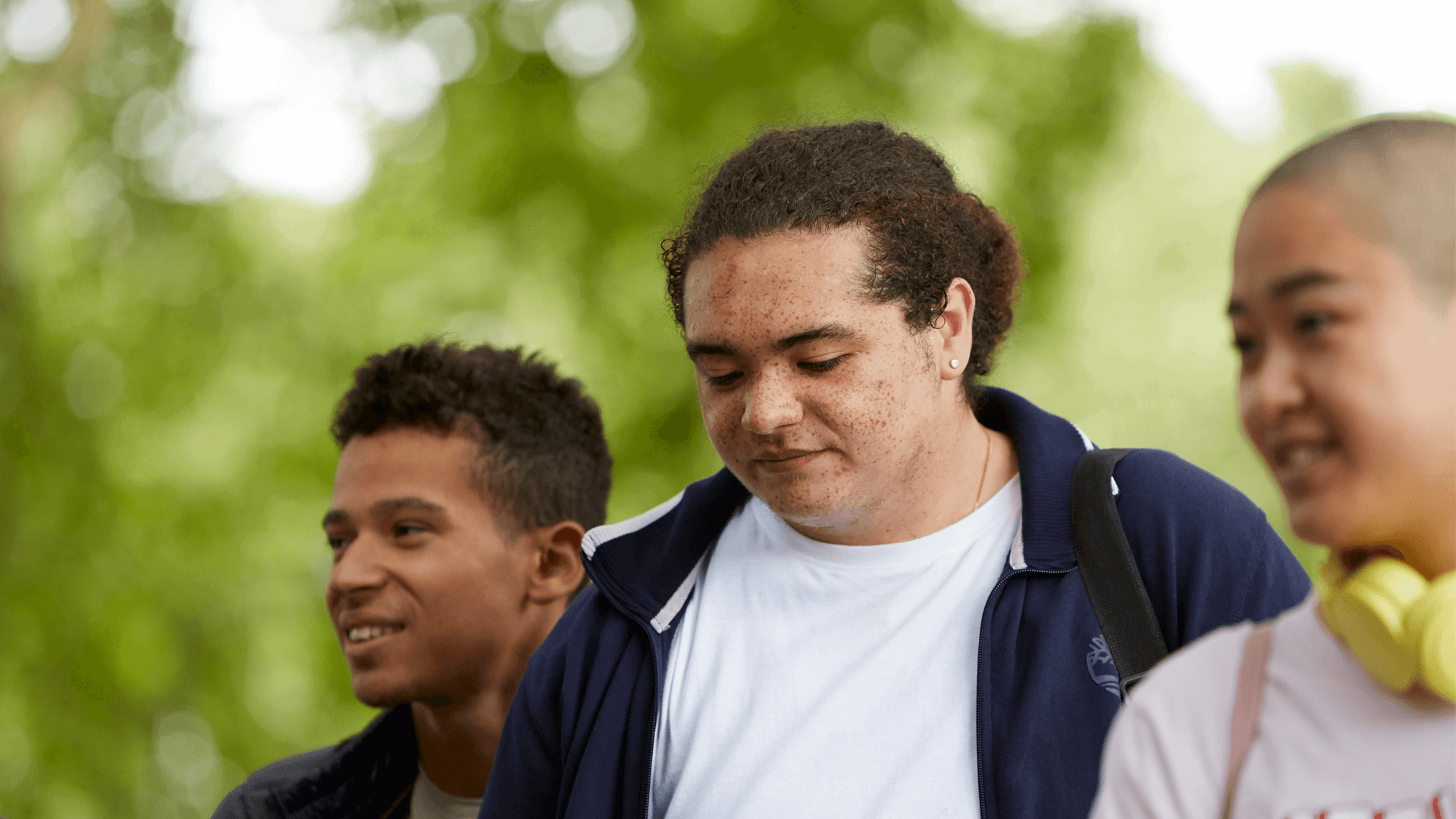 Three young people walking together through a park. Two of them are smiling while a boy in the centre looks worried.